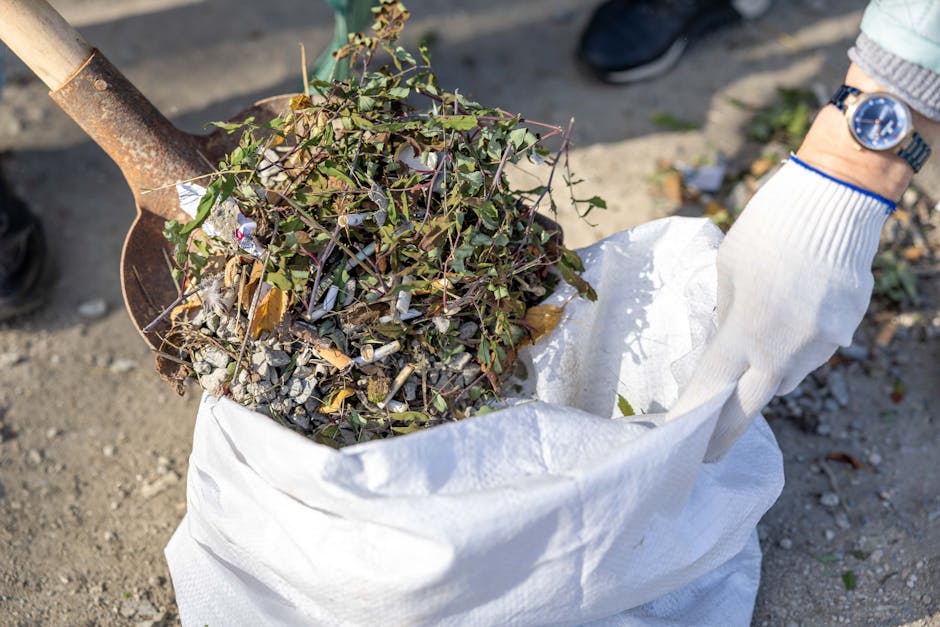 Close-up of a gardener wearing a white glove using a small trowel to scoop up a mix of garden debris including small branches, leaves, and gravel into a large white garden sack. The scene is outdoors,